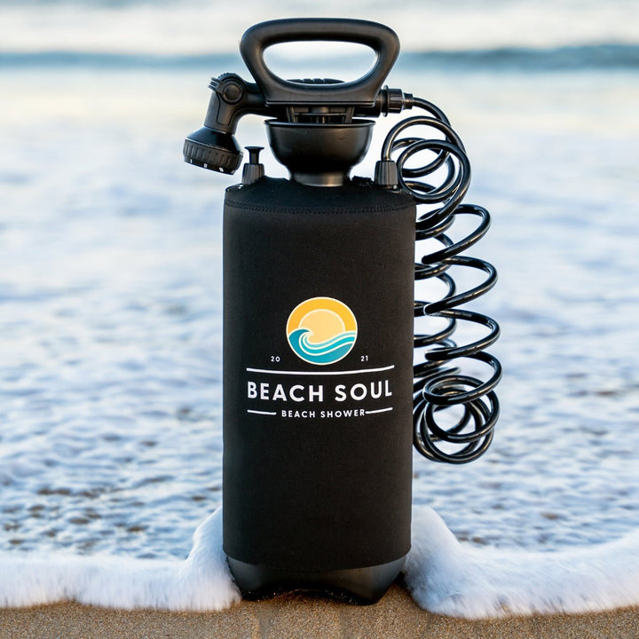 A black Beach Soul Beach Shower pump tank with a coiled hose and spray nozzle, standing on white foam at the edge of a beach with the ocean in the background.